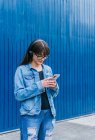 Mujer positiva en la navegación de auriculares en el teléfono inteligente mientras está de pie sobre fondo azul en la calle - foto de stock