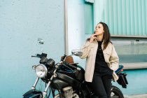 Уверенная женщина-байкер, опирающаяся на мотоцикл, припаркованный на обочине дороги в городе и курящая сигарету, отворачиваясь — стоковое фото
