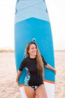 Счастливая девушка-серфингистка стоит с синей доской SUP на песчаном берегу летом и смотрит в сторону — стоковое фото