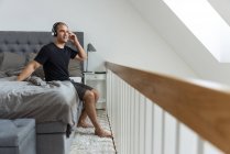 Vista lateral de un hombre tranquilo que se pone los auriculares y escucha música mientras está sentado en la cama después de despertar en el dormitorio y disfrutar de la mañana - foto de stock