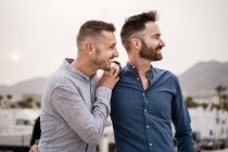 Пара гомосексуальних чоловіків у сорочках, які обіймаються, дивлячись на гавань проти океану і гори — стокове фото