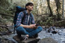 Vista laterale del contenuto escursionista maschio con zaino seduto vicino al lago nei boschi e la navigazione cellulare durante il viaggio — Foto stock