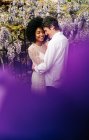 Vista lateral do casal multirracial amoroso abraçando no parque com flores de wisteria roxo florescendo no verão — Fotografia de Stock