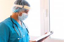 Focused male medic with stethoscope and in mask reading medical report on clipboard while standing in clinic — Stock Photo