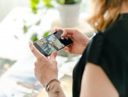 Alegre diseñadora femenina tomando fotos de pinturas en smartphone mientras está de pie cerca de la mesa y trabajando en un espacio de trabajo creativo - foto de stock