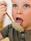 Adorable niño comiendo sopa de crema apetitosa y lamiendo cuchara durante el almuerzo en casa - foto de stock