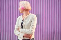 Mulher alternativa encantadora com cabelo tingido e em roupas da moda em pé contra a parede violeta na cidade e olhando para longe — Fotografia de Stock