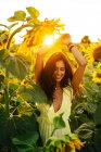 Graciosa feliz jovem hispânica fêmea em elegante vestido amarelo de pé com braços levantados em meio a girassóis florescendo no campo rural no dia ensolarado de verão com os olhos fechados — Fotografia de Stock