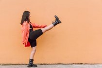 Visão lateral corpo cheio de alegre millennial feminino na roupa elegante chutando ar contra a parede laranja — Fotografia de Stock