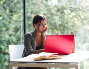 Seitenansicht einer Studentin, die am Netbook tippt, während sie mit Buch am Tisch sitzt und Hausaufgaben in der Universitätsbibliothek macht — Stockfoto