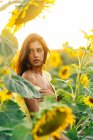 Вид сбоку на изящную молодую испаноязычную женщину в стильном желтом платье, стоящую посреди цветущих подсолнухов в сельской местности в солнечный летний день, отводя взгляд — стоковое фото