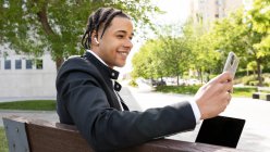 Vista lateral de un joven empresario étnico sonriente con trenzas haciendo videollamadas usando teléfonos inteligentes y auriculares TWS mientras está sentado en el banco con computadora portátil en la calle urbana - foto de stock