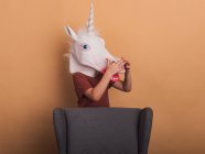 Anonymous kid in decorative unicorn mask with open mouth on beige background while touching his nose — Stock Photo