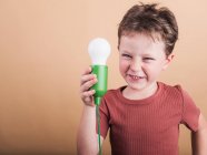 Pondering child in t shirt with plastic light bulb representing idea concept looking at camera on beige background — Stock Photo