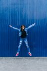 Hipster féminine excitée en tenue denim sautant avec les bras tendus sur fond bleu en ville et regardant la caméra — Photo de stock