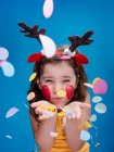 Girl with red painted cheeks and in antler headband blowing off vivid paper confetti while standing on blue background — Stock Photo