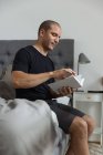 Vue latérale d'un mâle coûteux assis sur un lit doux le matin et lisant une histoire intéressante dans un livre après son réveil — Photo de stock