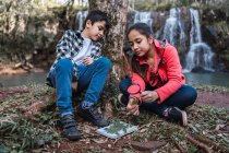 Ethnic girl with brother studying plant foliage with magnifying glass while sitting against river and cascade — Stock Photo