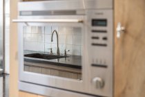 Torneira de metal e pia undermount refletindo na porta de vidro de construído no forno na cozinha contemporânea em apartamento — Fotografia de Stock