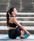 Young female athlete in black sportswear listening to music in true wireless earphones and doing warm up exercise while sitting on steps on city street — Stock Photo