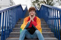 Знизу стильний гей-чоловік з ЛГБТ-прапором на плечах сидить на металевих сходах у місті і дивиться на камеру. — стокове фото