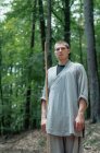 Hombre con palo sosteniendo la mano cerca del pecho mientras practica kung fu en el bosque mirando a la cámara - foto de stock
