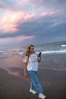 Стильная курчавая женщина, прогуливающаяся по пляжу у моря летним вечером и просматривающая мобильный телефон — стоковое фото
