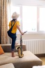 Back view of anonymous thoughtful barefoot child in hat with saxophone standing on sofa against window at home — Stock Photo