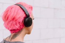 Vista lateral de una joven mujer anónima con pelo rosa brillante escuchando música con auriculares mientras está de pie cerca de la pared blanca - foto de stock