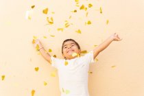 Happy boy in white t shirt standing with raised arms while tossing up pile of yellow hear confetti on light orange wall — Stock Photo