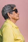 Vista laterale di felice donna moderna invecchiata con i capelli grigi e in occhiali da sole alla moda su sfondo rosa in studio e guardando altrove — Foto stock