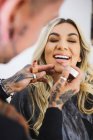 Mulher loura alegre sorrindo com olhos fechados enquanto artista de maquiagem tatuada manchando batom líquido nos lábios do modelo — Fotografia de Stock