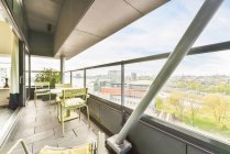 Стол со стульями расположен на просторном балконе современного жилого здания со стеклянным забором, смотрящим на городской пейзаж в летний день — стоковое фото