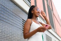 Bajo ángulo de sonriente mujer afroamericana con brads mensajería en las redes sociales a través del teléfono móvil mientras está de pie en la calle con edificios antiguos en Barcelona - foto de stock