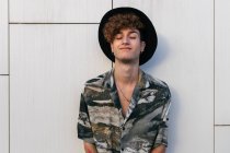 Молодой тщеславный человек в стильной одежде в шляпе, стоящий на черепичной стене, глядя в сторону — стоковое фото