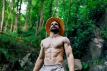 From below of fit shirtless male traveler with backpack and in hat standing in rocky woods and looking away — Stock Photo