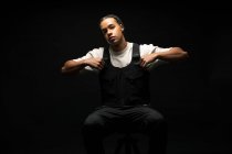Bonito jovem macho étnico com tranças afro vestido com roupas pretas e brancas olhando para a câmera enquanto sentado no estúdio escuro — Fotografia de Stock