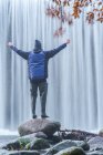 Вид на путешествующего мужчину в теплой одежде, стоящего на валуне с протянутыми руками и восхищающимся видом на быстрый водопад на реке Лозоя в Национальном парке Гуадарама — стоковое фото