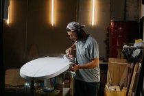 Male shaper using electric planer and polishing surface of surfboard in workshop — Stock Photo