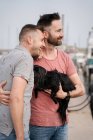 Вид сбоку веселых взрослых гомосексуальных мужчин с симпатичной собакой, смотрящих в гавань — стоковое фото