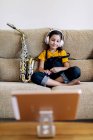 Запоминающийся ребенок в наушниках и саксофоне на диване, записывающий дома — стоковое фото