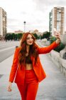 Deliciosa hembra de moda con cabello rojo y traje naranja caminando por la calle en la ciudad y tomando autorretrato en el teléfono inteligente - foto de stock