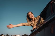 Bajo ángulo de mujer alegre con el brazo extendido que sobresale de la ventana del coche y disfruta de la libertad en la noche de verano - foto de stock
