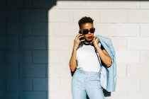 Stilvolle erwachsene Afroamerikanerin mit modernem Haarschnitt und Jacke, die mit dem Handy gegen eine geflieste Wand mit Schatten im Sonnenlicht spricht — Stockfoto