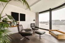 Fragment of interior design of modern luxury apartment with comfortable leather armchairs and foot stool placed in front of panoramic windows overlooking channel in city — Stock Photo
