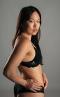 Fiduciosa donna asiatica in lingerie nera in piedi su sfondo grigio in studio e guardando la fotocamera — Foto stock