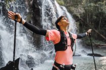 Senderista masculino soñador con bastones de trekking en manos extendidas de pie con los ojos cerrados cerca de la cascada en el bosque y disfrutando de la libertad - foto de stock