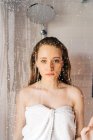 Femme enveloppée dans une serviette douce blanche debout derrière la porte en verre humide de la cabine de douche et regardant la caméra — Photo de stock