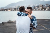 Uomo sincero che abbraccia partner omosessuale irriconoscibile guardando lontano contro il lago e il monte in città — Foto stock