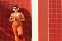 Весела етнічна жінка-спортсменка з пишним тілом і мобільним телефоном слухає пісню з навушників, дивлячись в сторону плиткової стіни — стокове фото
